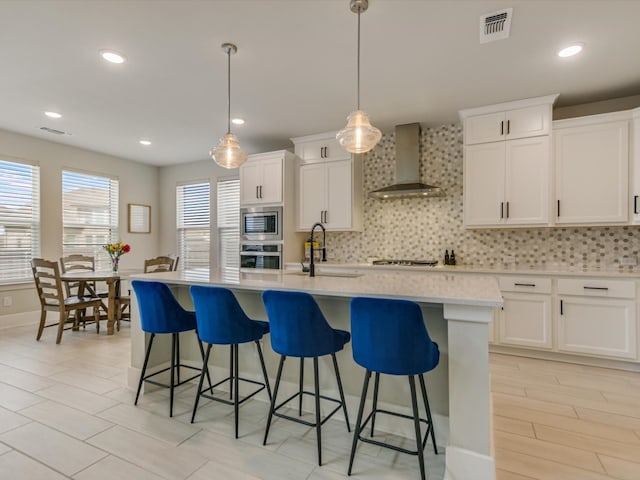 kitchen featuring white cabinetry, stainless steel appliances, wall chimney range hood, an island with sink, and decorative light fixtures