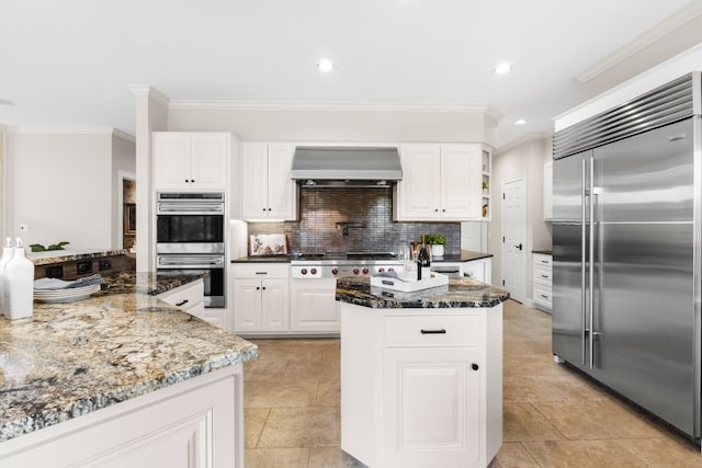 kitchen featuring wall chimney exhaust hood, white cabinetry, stainless steel appliances, dark stone countertops, and crown molding