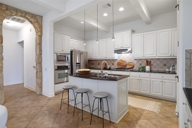 kitchen featuring beamed ceiling, appliances with stainless steel finishes, a center island with sink, and white cabinetry