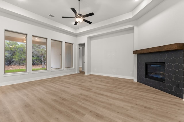 unfurnished living room featuring a tiled fireplace, ceiling fan, light wood-type flooring, and a tray ceiling