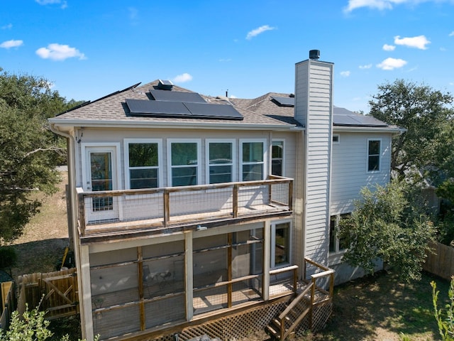 back of property with roof mounted solar panels, a shingled roof, a chimney, and a balcony