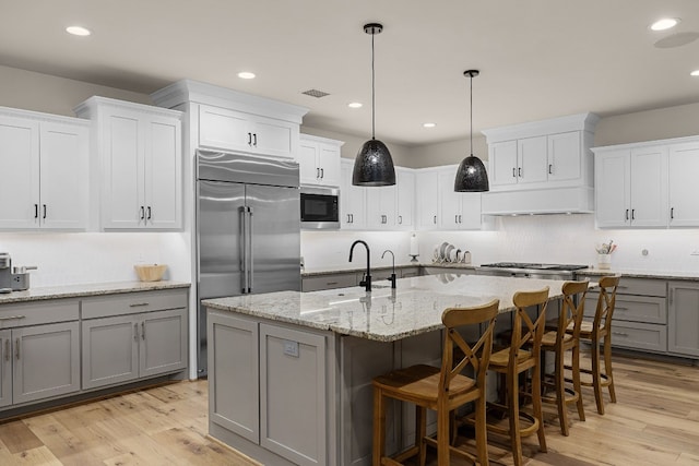 kitchen featuring light wood-type flooring, built in appliances, hanging light fixtures, gray cabinetry, and a center island with sink