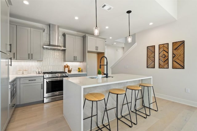 kitchen with wall chimney range hood, sink, light hardwood / wood-style flooring, an island with sink, and appliances with stainless steel finishes