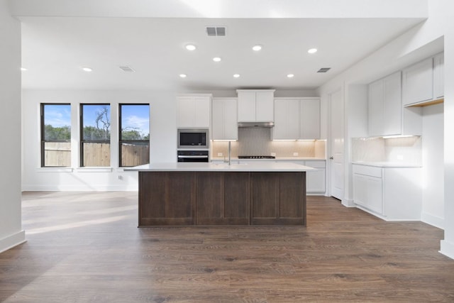 kitchen with oven, built in microwave, dark wood-style floors, tasteful backsplash, and an island with sink