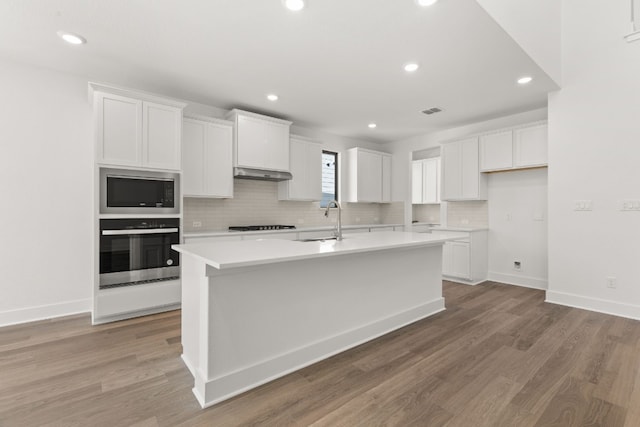 kitchen featuring built in microwave, sink, a center island with sink, oven, and white cabinets