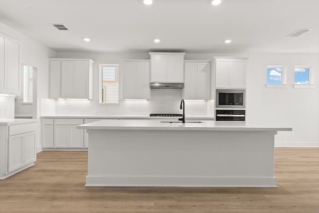 kitchen with a kitchen island with sink, oven, white cabinets, and light wood-type flooring