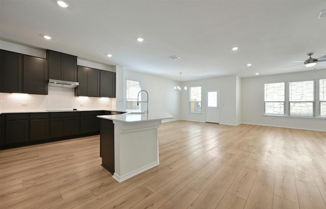 kitchen featuring an island with sink, light wood-type flooring, decorative backsplash, sink, and ceiling fan with notable chandelier