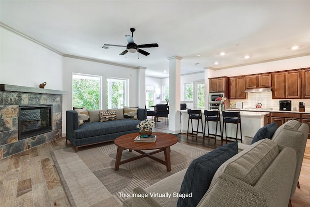 living room with a tile fireplace, a ceiling fan, ornamental molding, light wood finished floors, and decorative columns