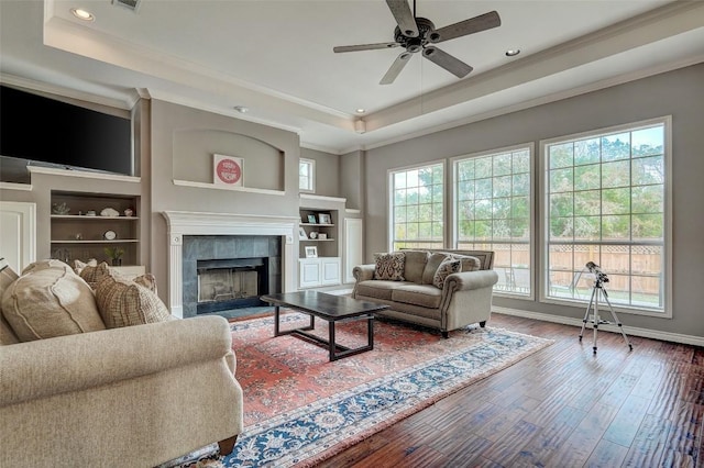 living room with crown molding, a fireplace, wood-type flooring, built in shelves, and a raised ceiling