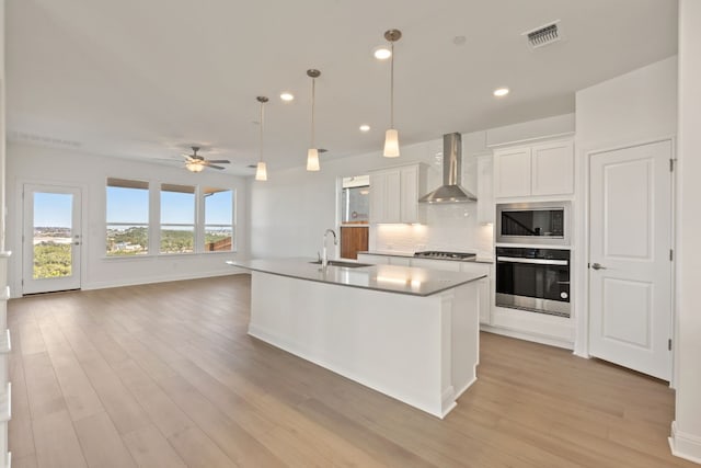 kitchen with sink, wall chimney exhaust hood, stainless steel appliances, a kitchen island with sink, and white cabinets