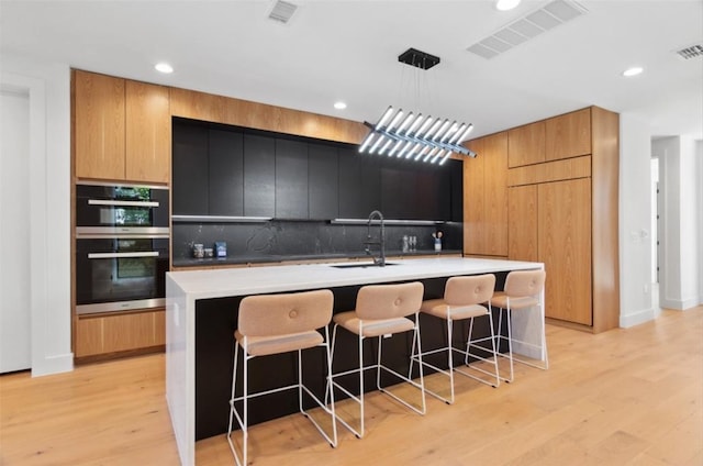 kitchen with backsplash, stainless steel double oven, light hardwood / wood-style flooring, a center island with sink, and sink