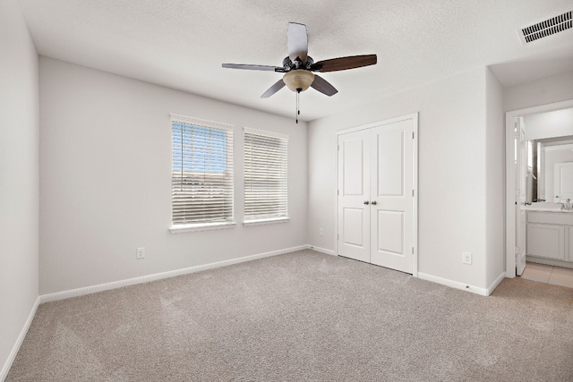 unfurnished bedroom featuring a closet, a textured ceiling, light carpet, ensuite bathroom, and ceiling fan