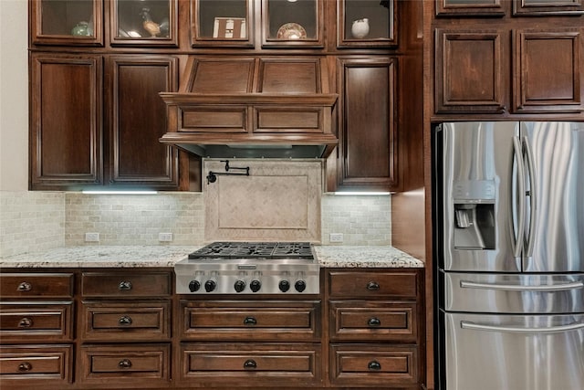 kitchen featuring stainless steel appliances, range hood, light stone countertops, tasteful backsplash, and glass insert cabinets