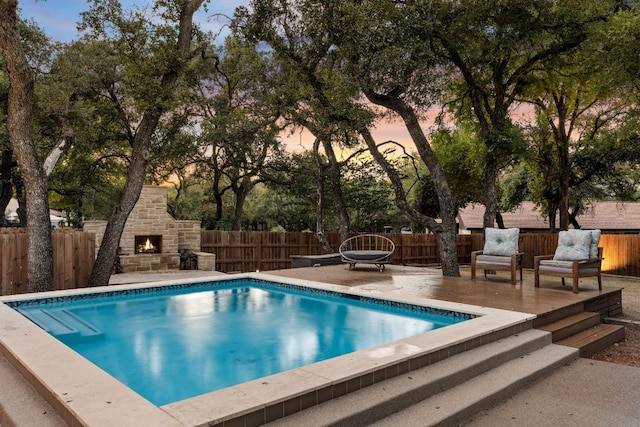 pool at dusk with an outdoor stone fireplace and a deck