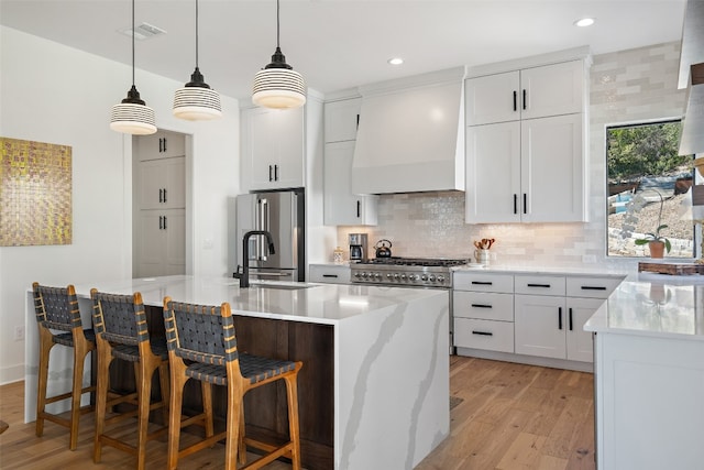 kitchen with high end fridge, custom exhaust hood, a kitchen island with sink, and white cabinetry