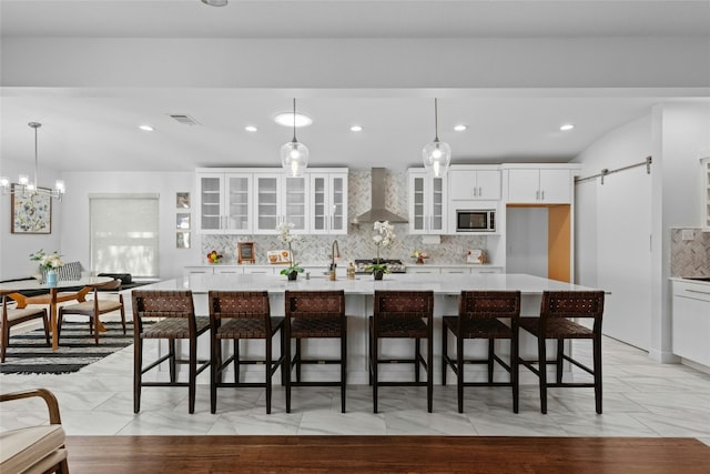 kitchen featuring stainless steel microwave, wall chimney range hood, a barn door, a kitchen bar, and white cabinets