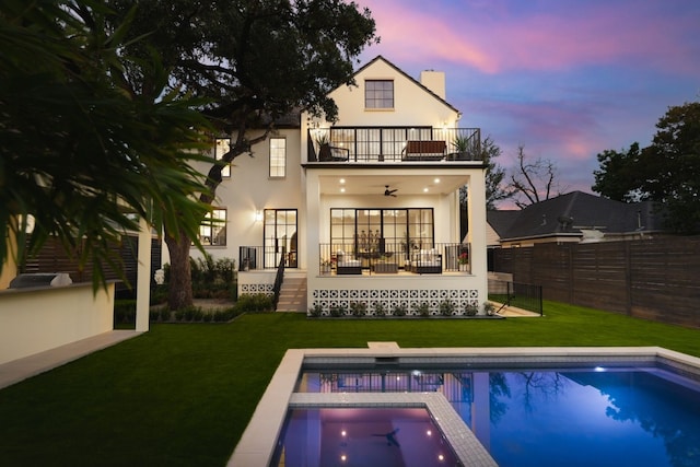 back of property at dusk featuring a lawn, a balcony, ceiling fan, fence, and stucco siding