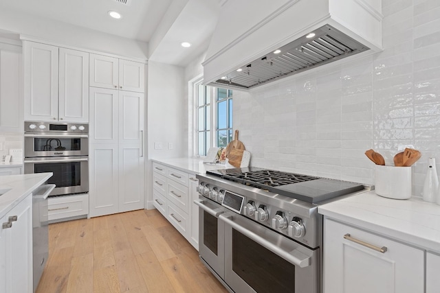 kitchen with light hardwood / wood-style floors, tasteful backsplash, white cabinetry, appliances with stainless steel finishes, and premium range hood