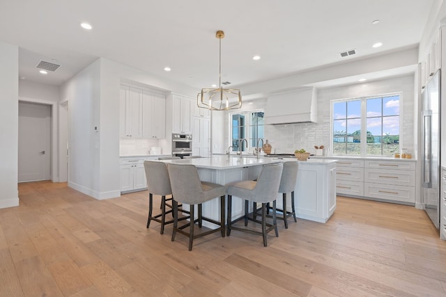 kitchen featuring light hardwood / wood-style floors, white cabinets, custom exhaust hood, decorative light fixtures, and a center island with sink