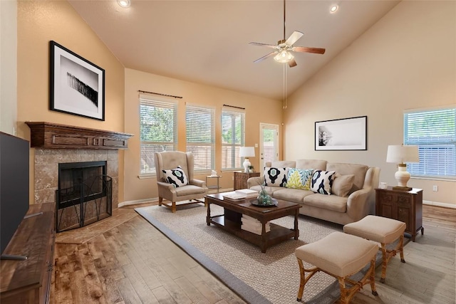 living room featuring a tiled fireplace, a ceiling fan, high vaulted ceiling, light wood-type flooring, and baseboards