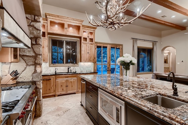 kitchen featuring beamed ceiling, sink, dark stone counters, wall chimney range hood, and appliances with stainless steel finishes