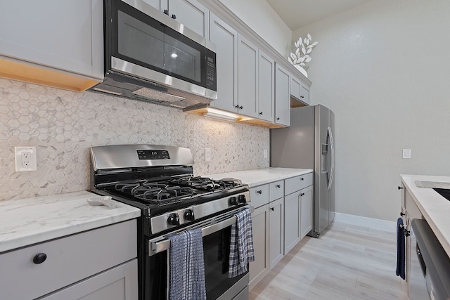 kitchen with gray cabinets, appliances with stainless steel finishes, decorative backsplash, light hardwood / wood-style floors, and light stone countertops