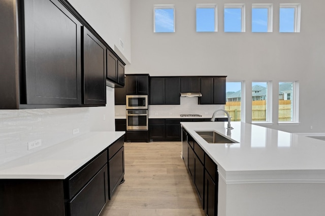 kitchen with appliances with stainless steel finishes, a towering ceiling, an island with sink, sink, and light hardwood / wood-style flooring