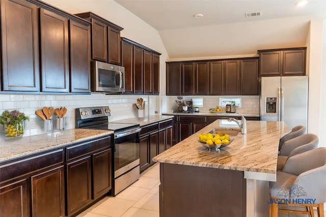 kitchen featuring backsplash, vaulted ceiling, a center island with sink, light tile patterned flooring, and appliances with stainless steel finishes