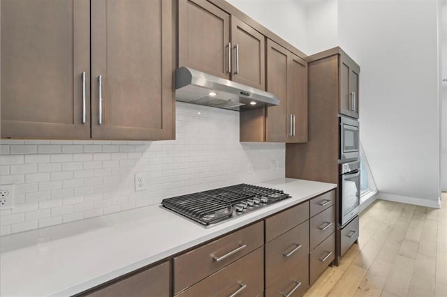 kitchen with under cabinet range hood, light countertops, appliances with stainless steel finishes, light wood-type flooring, and decorative backsplash