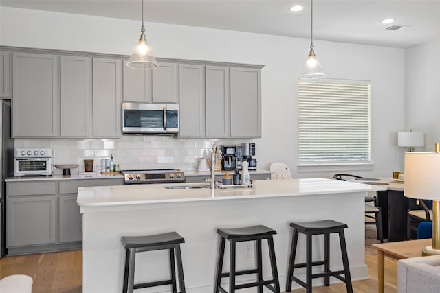 kitchen featuring appliances with stainless steel finishes, light hardwood / wood-style floors, backsplash, gray cabinets, and a center island with sink