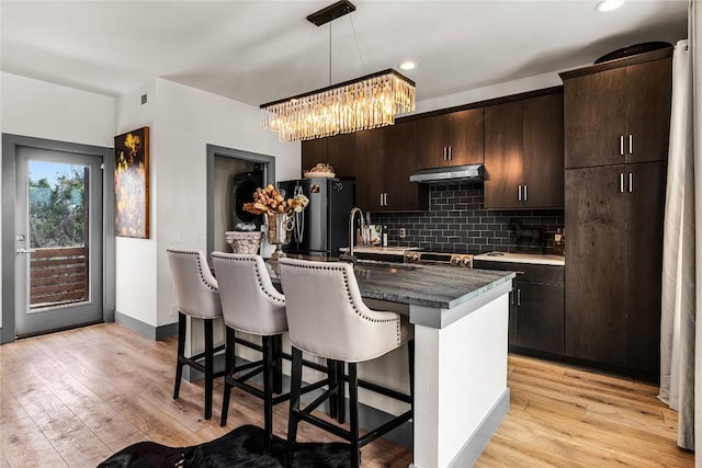 kitchen featuring a kitchen island with sink, hanging light fixtures, stainless steel fridge, light hardwood / wood-style floors, and dark brown cabinetry