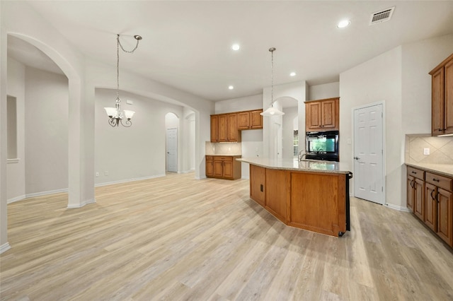 kitchen with light stone counters, an island with sink, hanging light fixtures, an inviting chandelier, and light hardwood / wood-style floors