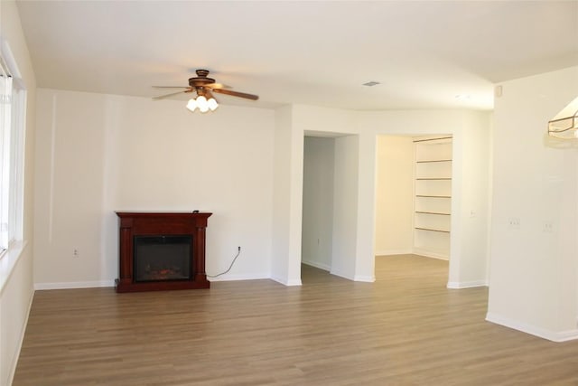 unfurnished living room featuring a glass covered fireplace, ceiling fan, baseboards, and wood finished floors