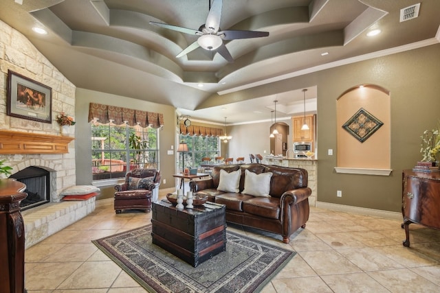 tiled living room with a raised ceiling, a stone fireplace, crown molding, and ceiling fan