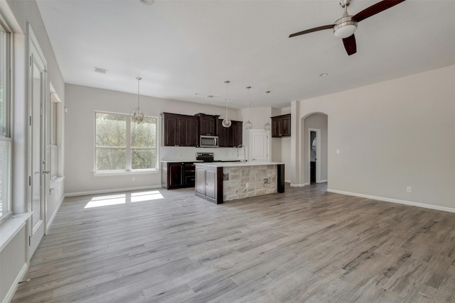 kitchen with dark brown cabinets, light hardwood / wood-style floors, an island with sink, hanging light fixtures, and appliances with stainless steel finishes