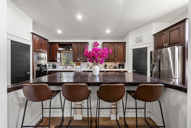 kitchen with a textured ceiling, appliances with stainless steel finishes, a kitchen bar, dark hardwood / wood-style flooring, and dark brown cabinets