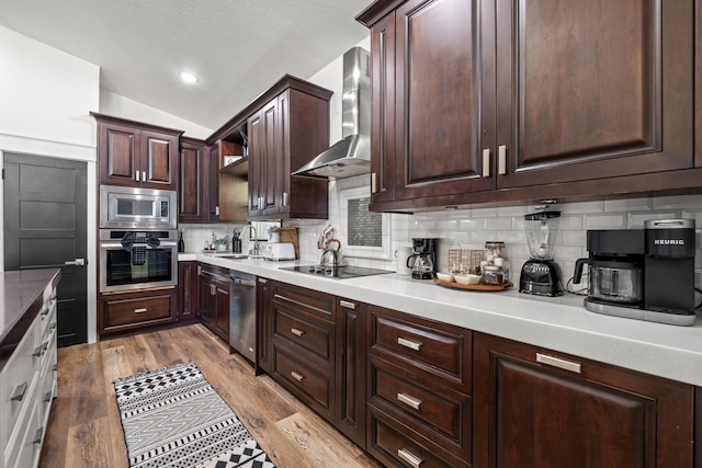 kitchen with appliances with stainless steel finishes, decorative backsplash, wall chimney range hood, light hardwood / wood-style flooring, and dark brown cabinetry