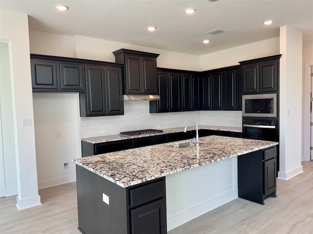 kitchen with appliances with stainless steel finishes, light wood-type flooring, tasteful backsplash, sink, and an island with sink