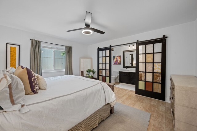 bedroom with light wood-type flooring, ceiling fan, ensuite bathroom, and a barn door