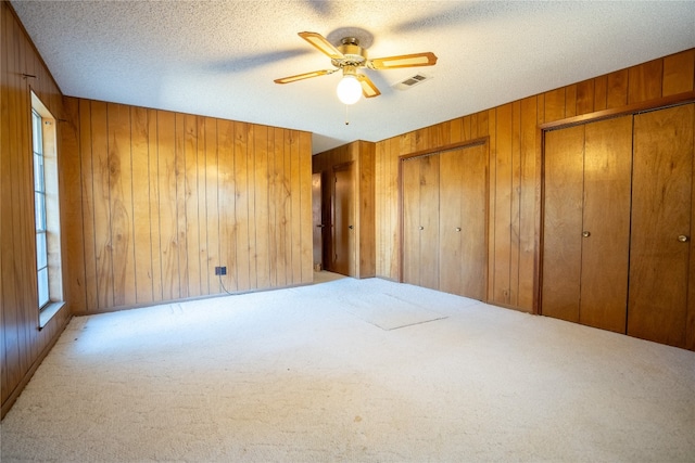 unfurnished bedroom featuring a textured ceiling, multiple closets, light colored carpet, wooden walls, and ceiling fan