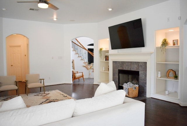 living room with built in shelves, ceiling fan, dark wood-type flooring, and a tiled fireplace