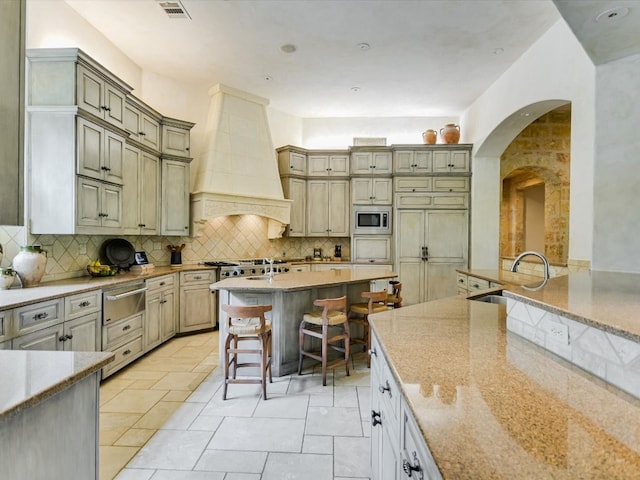 kitchen with a kitchen bar, backsplash, appliances with stainless steel finishes, light stone countertops, and premium range hood