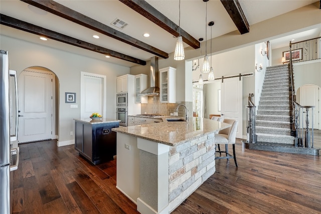 kitchen featuring appliances with stainless steel finishes, hanging light fixtures, light stone counters, a barn door, and wall chimney exhaust hood