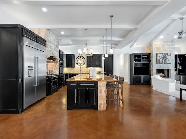 kitchen with decorative backsplash, light stone counters, decorative light fixtures, a kitchen island with sink, and stainless steel built in fridge