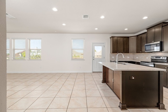 kitchen with light stone counters, tasteful backsplash, dark brown cabinets, a center island with sink, and appliances with stainless steel finishes