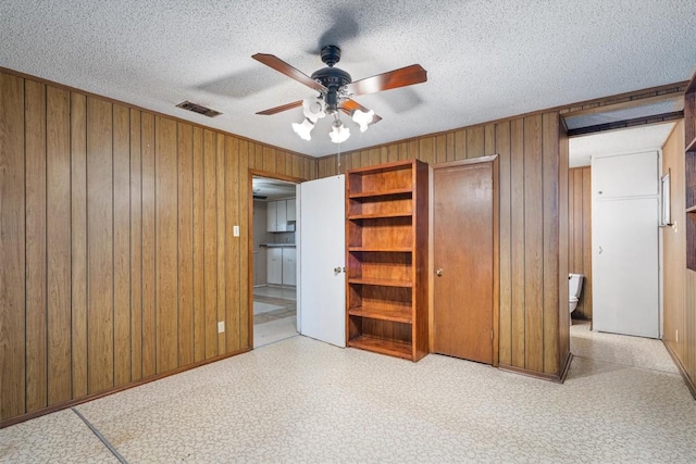 unfurnished bedroom featuring connected bathroom, ceiling fan, wooden walls, and a textured ceiling