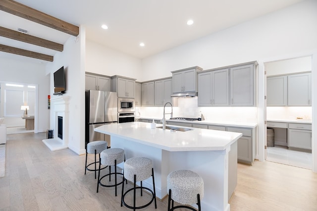 kitchen with light wood-type flooring, beamed ceiling, a kitchen island with sink, sink, and appliances with stainless steel finishes