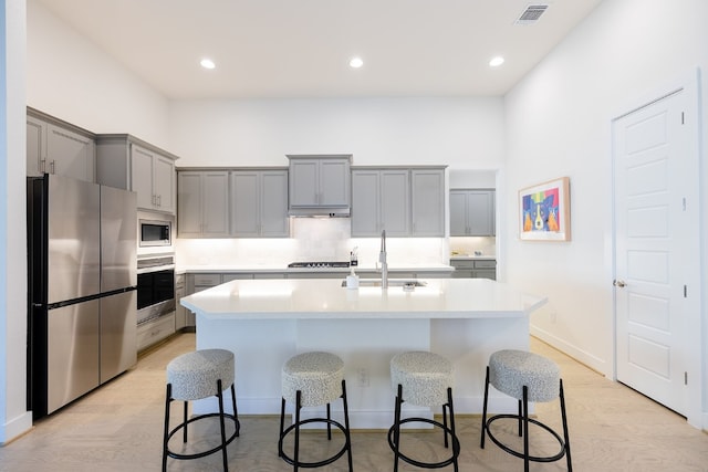 kitchen featuring appliances with stainless steel finishes, gray cabinetry, an island with sink, light wood-type flooring, and sink