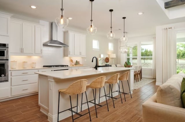 kitchen featuring hanging light fixtures, wall chimney exhaust hood, a kitchen island with sink, and white cabinetry