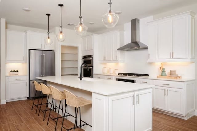 kitchen featuring an island with sink, light wood-type flooring, stainless steel appliances, white cabinets, and wall chimney exhaust hood